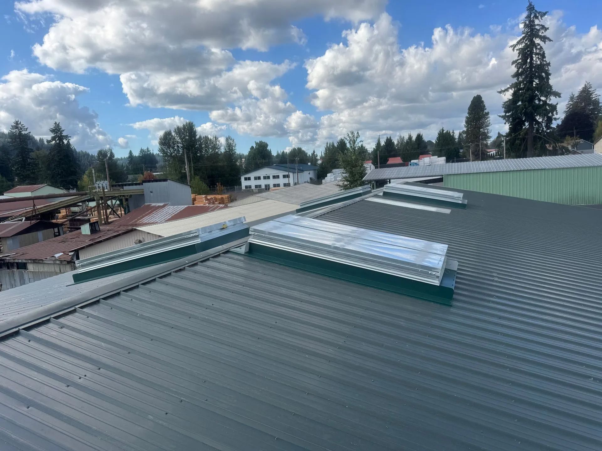Rooftop with gray metal panels, skylights, and a cloudy sky over nearby buildings and trees