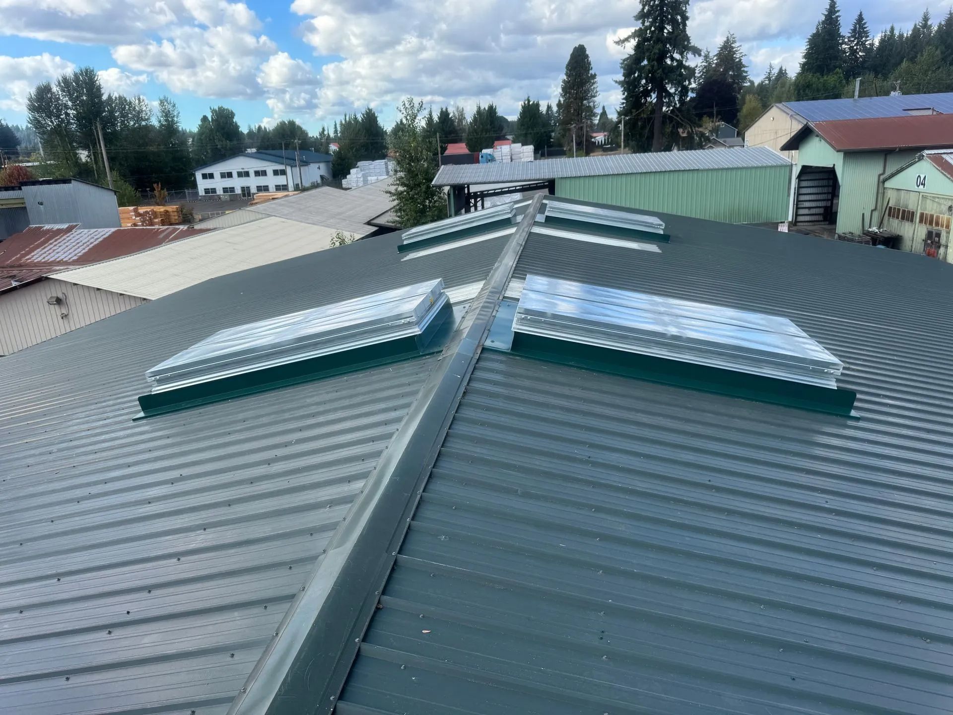 Rooftop with green metal panels and skylight windows above a neighborhood under a cloudy sky