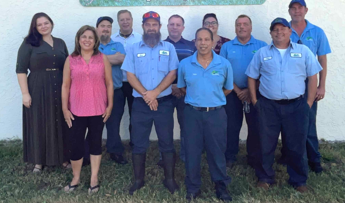 Group of people posing outdoors in front of a white wall, wearing blue shirts and dark uniforms.