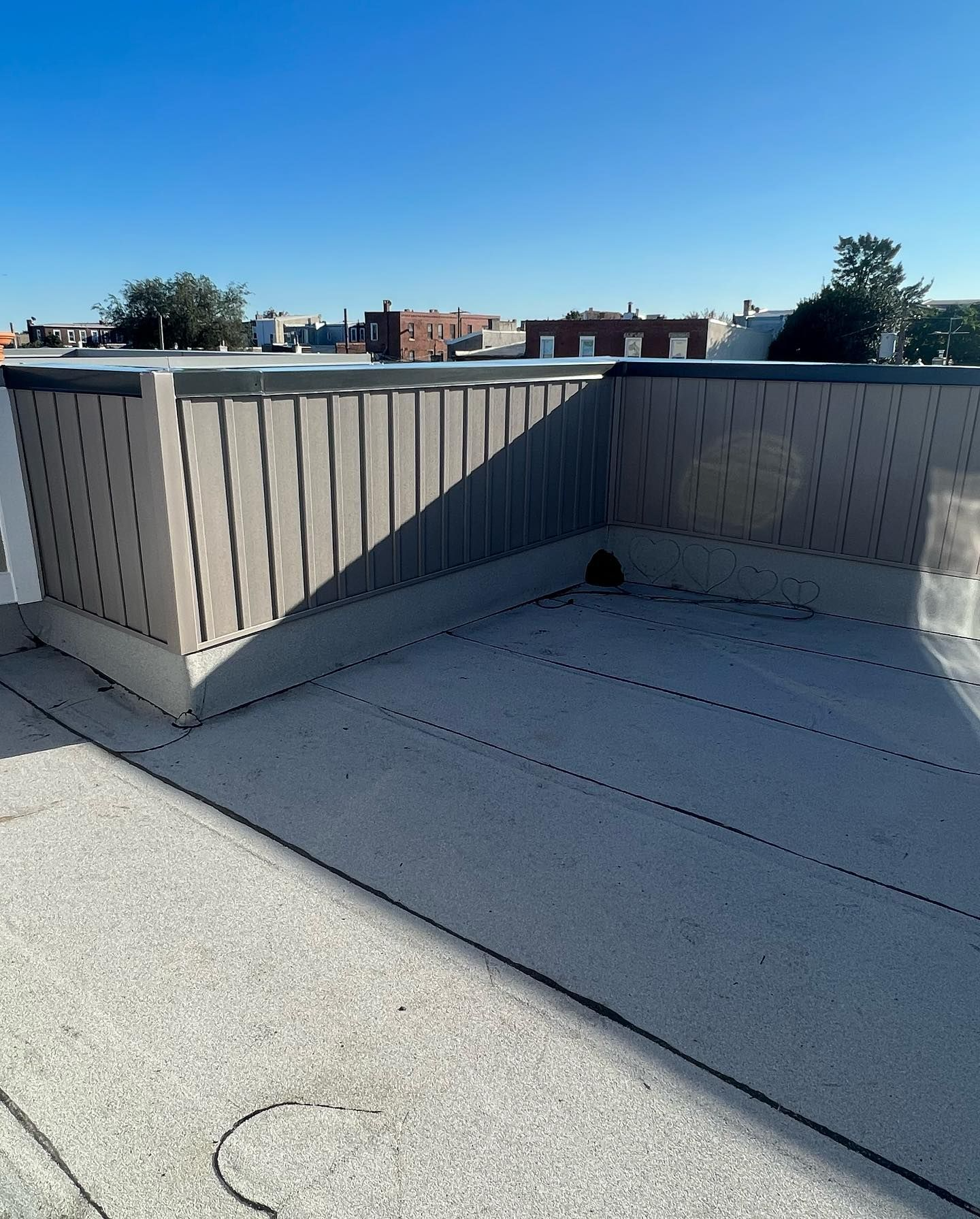 Rooftop with a low fence. Gray flat roof, light gray fence, buildings in the background under blue sky.