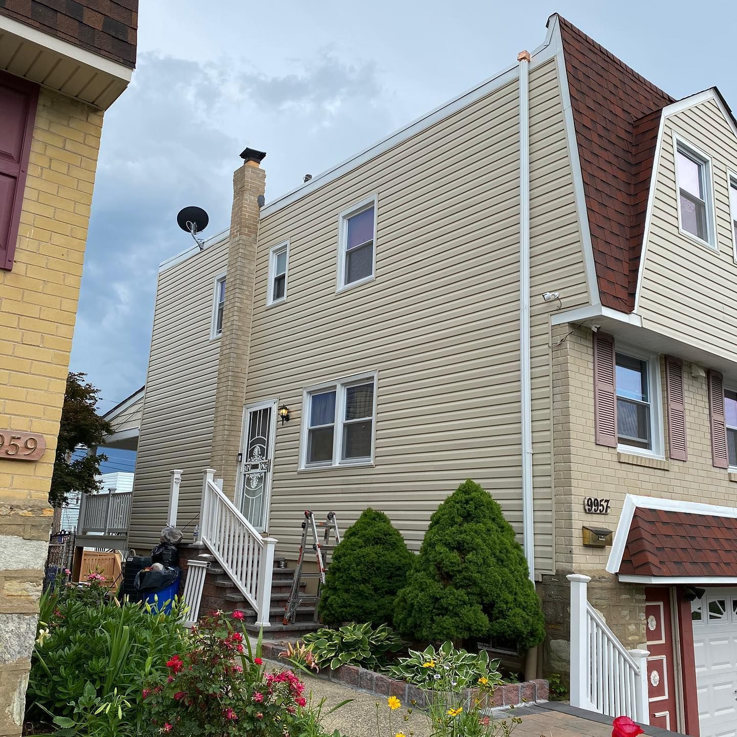 Two-story house with tan siding, brown roof, white railing, and small front yard with bushes.