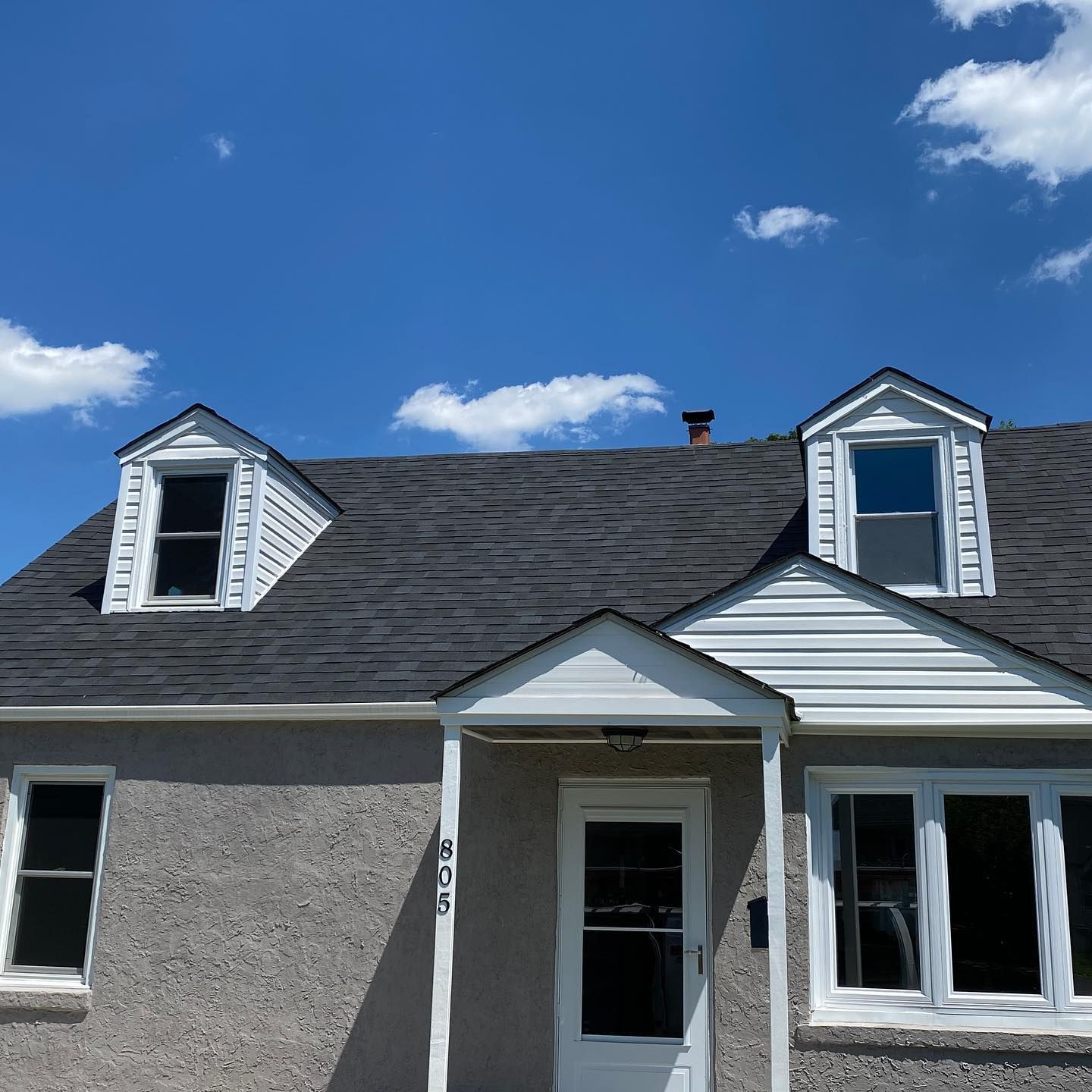 Gray stucco house with two dormers, white trim, and a blue sky with clouds.