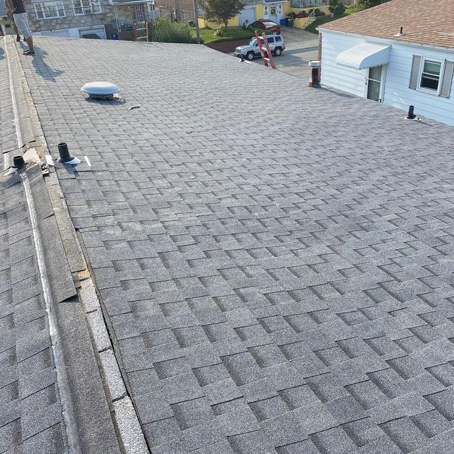 Flat shingled roof, light gray, with multiple vents. Houses in background, sunny day.