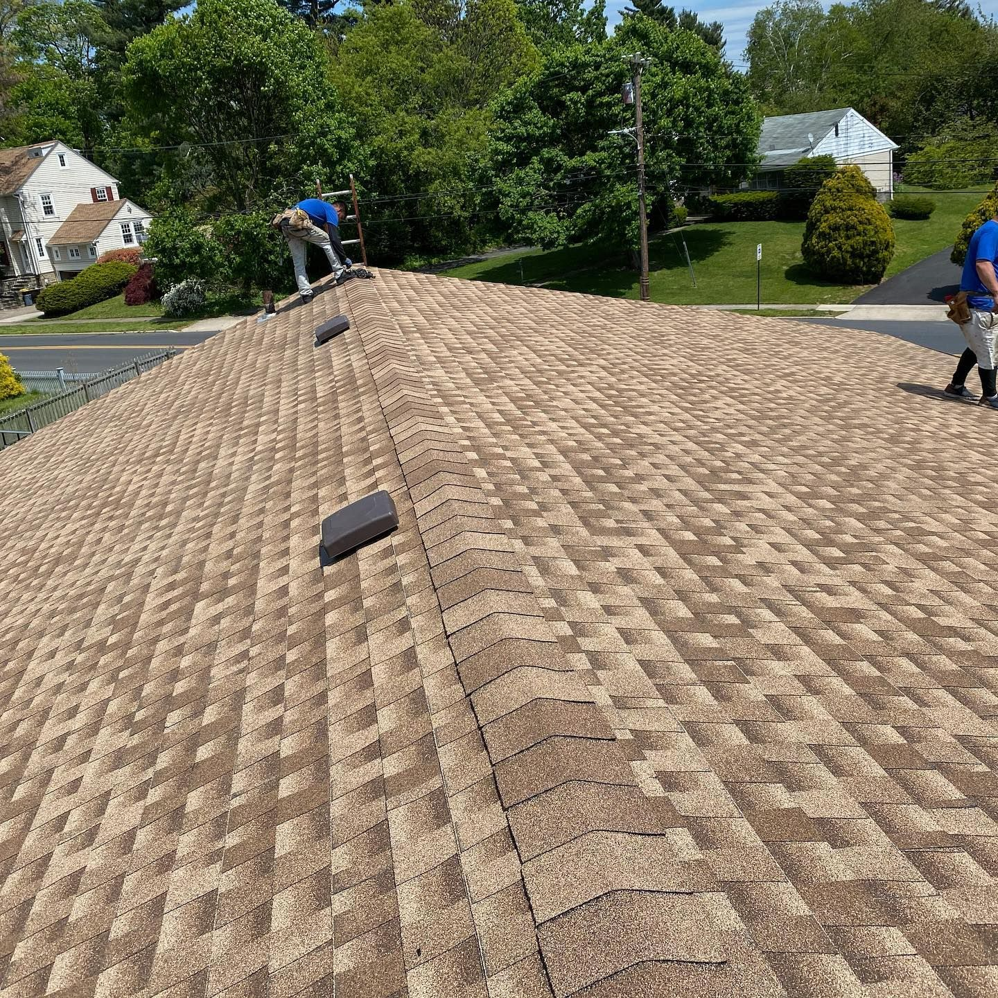 Workers on a tan shingle roof under a blue sky, several houses in the background.
