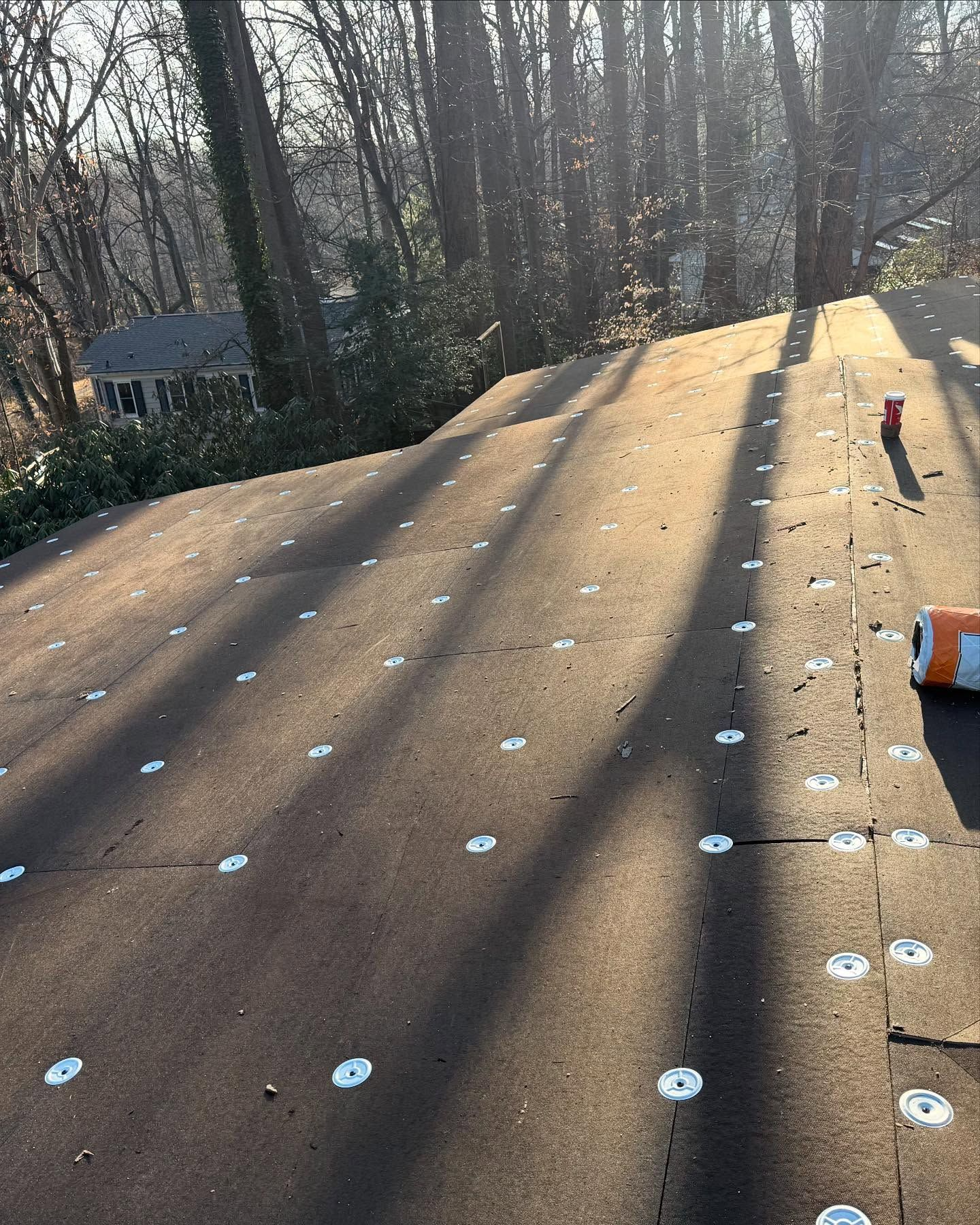 Roof covered in brown underlayment, with metal fasteners. Trees and a house are in the background.