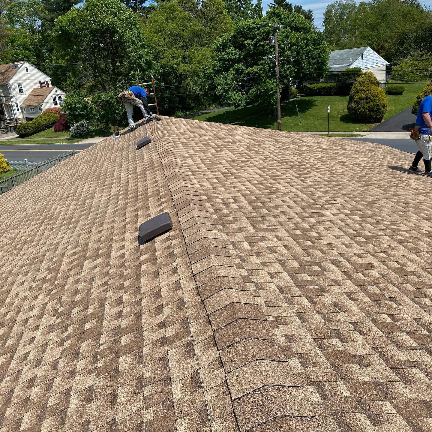 Workers on a brown shingled roof, trees and houses in the background. Sunny day.