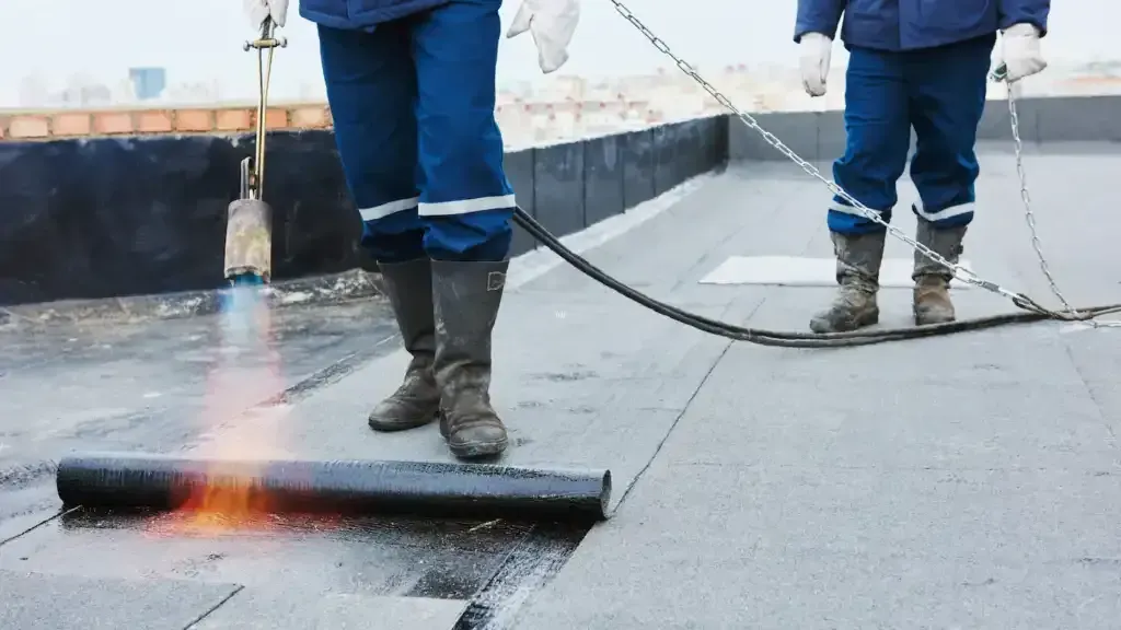 Two workers on a flat roof, applying roofing material with a torch.