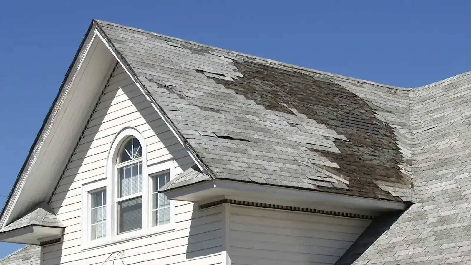 Damaged house roof with missing and discolored shingles against a clear blue sky.