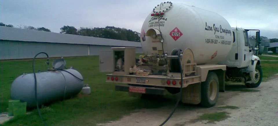 A white propane truck is refueling a gray propane tank on a grassy area near a building.