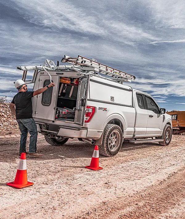 Worker loading gear into a white pickup truck on a dirt road, with orange cones under a cloudy sky