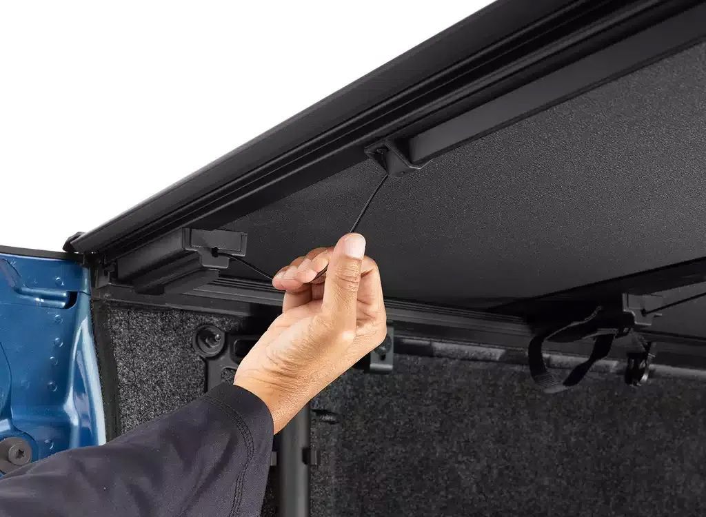 Hand adjusting a latch on the inside of a black truck bed cover near a blue vehicle panel