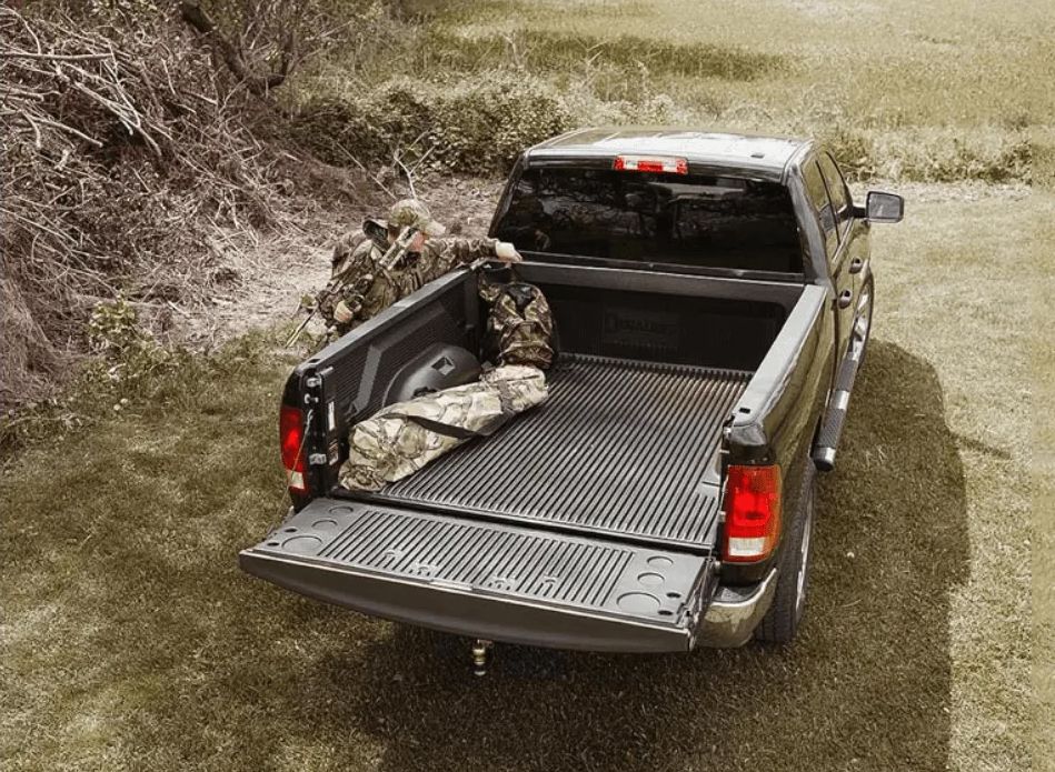 Pickup truck bed with an open tailgate and camouflage gear parked on a dirt road in a wooded area