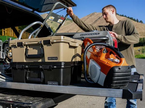 Man loading tan and black storage boxes and an orange chainsaw onto a trailer rack