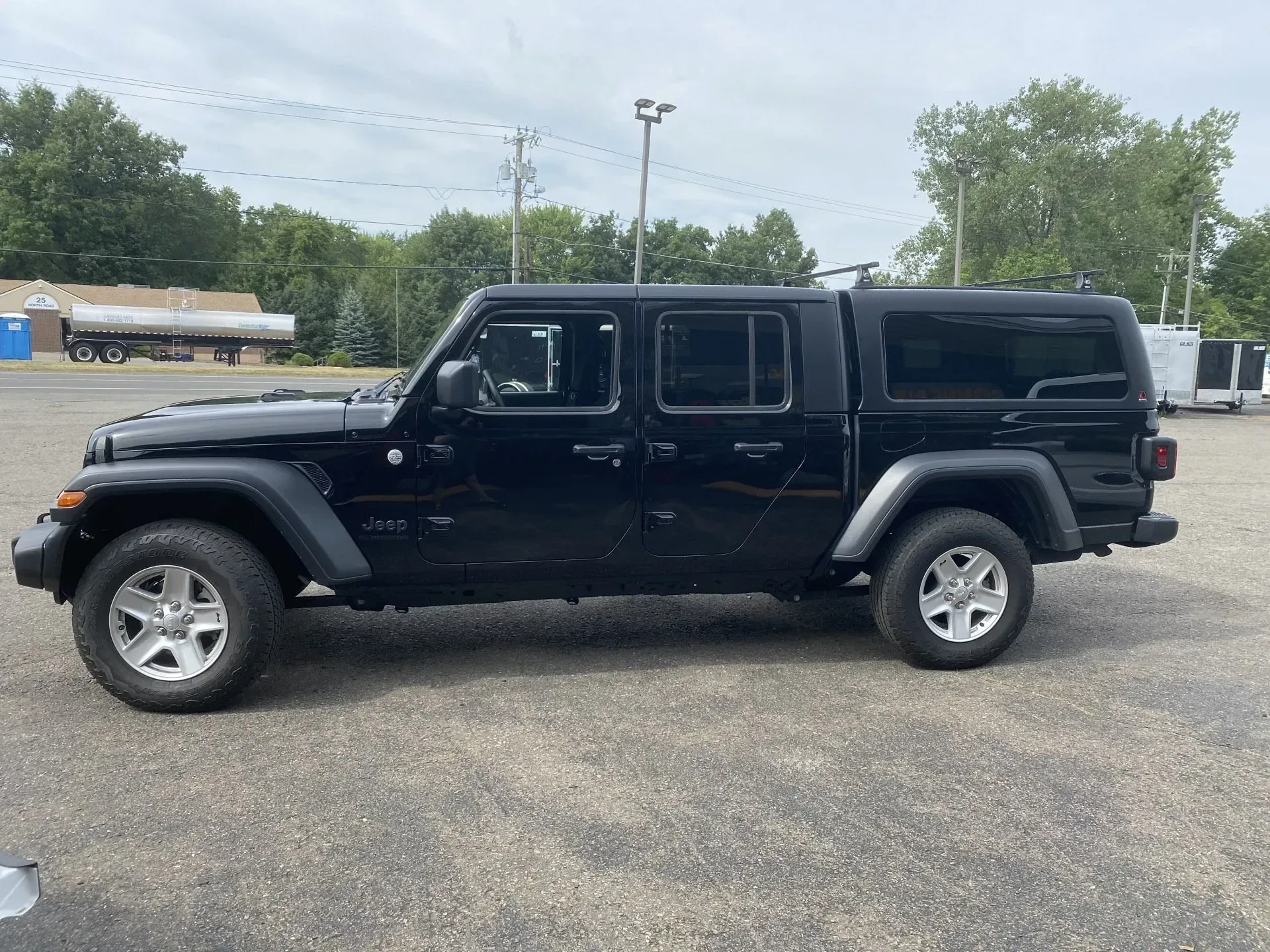 Black Jeep Gladiator pickup parked on gravel lot, side view with trees and sky in background