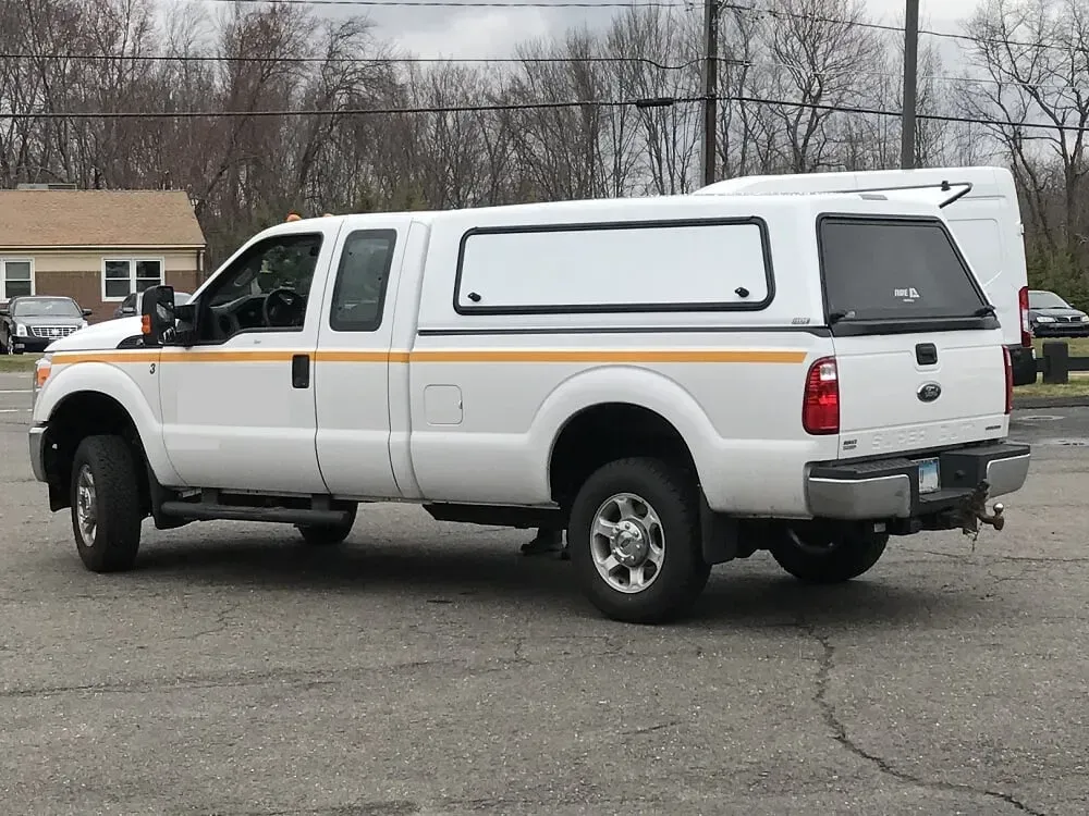 White pickup truck with camper shell parked on an asphalt lot near trees and houses