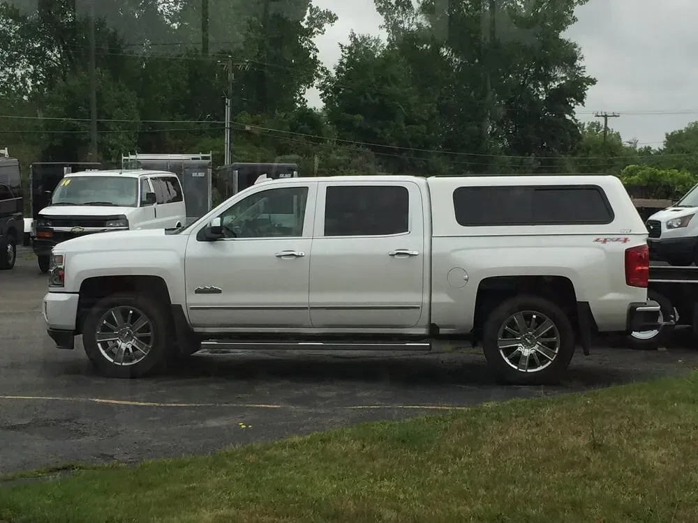 White pickup truck with canopy parked in a wet lot near trees and other vehicles