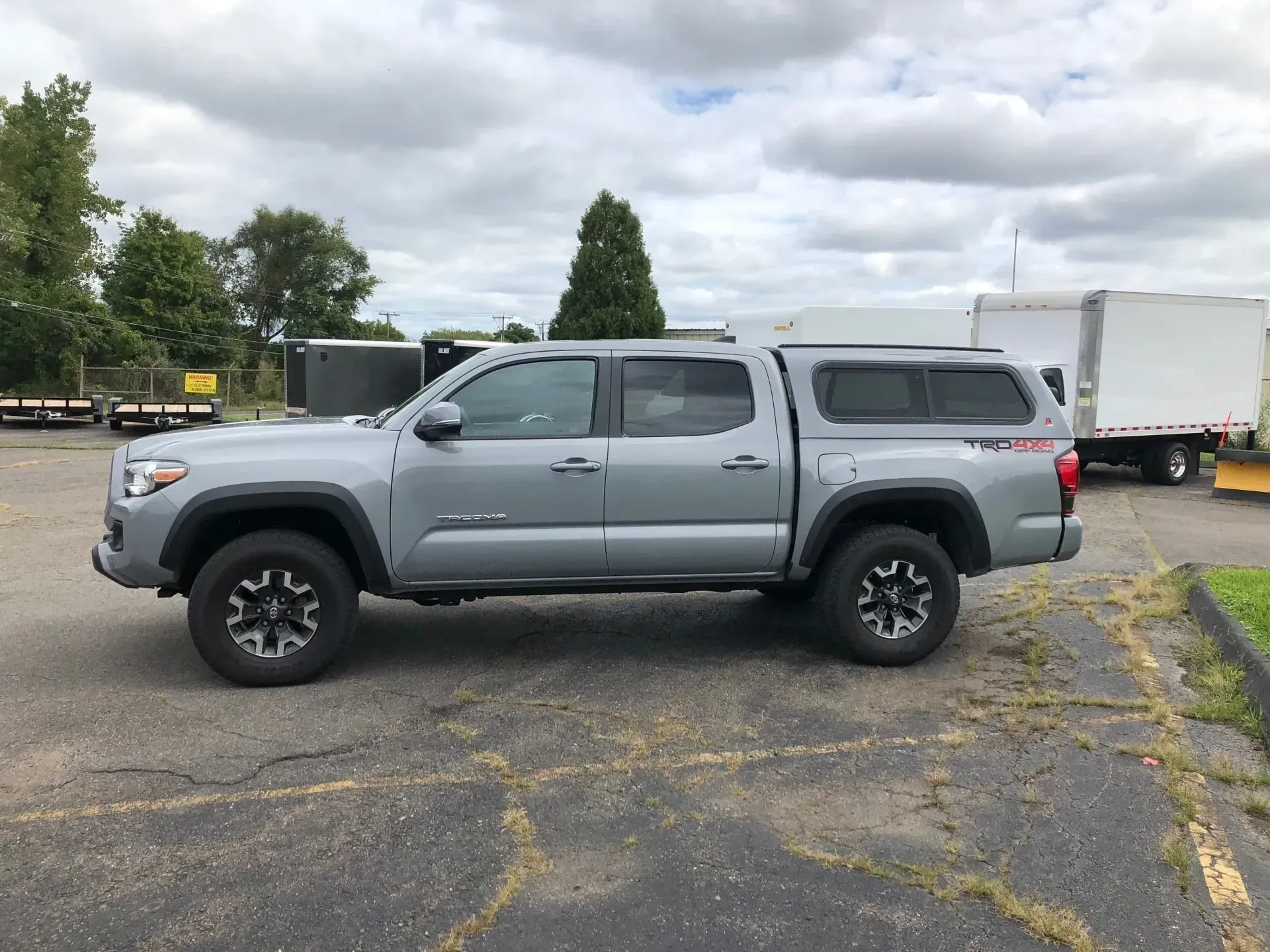 Gray pickup truck parked in a lot beside a white box truck under cloudy skies