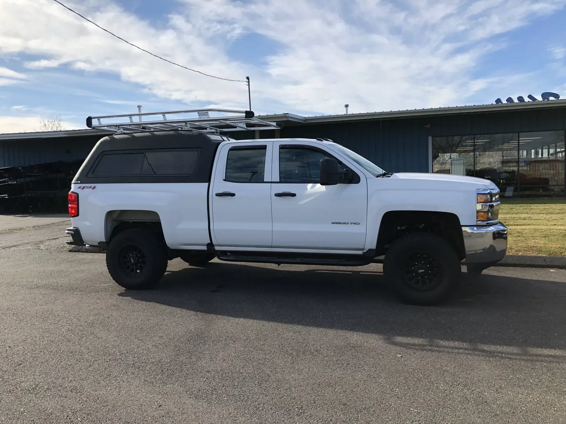 White pickup truck with camper shell and roof rack parked on a road in front of a building.