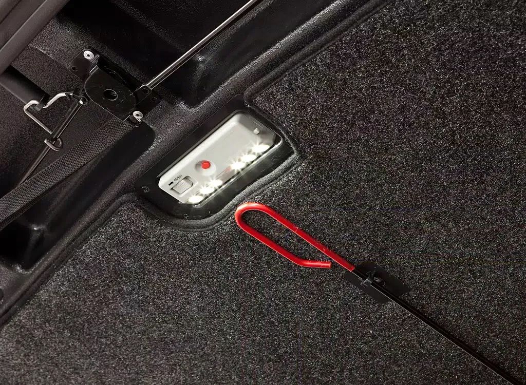 Car trunk with a small silver device and a red looped strap on dark carpet flooring.