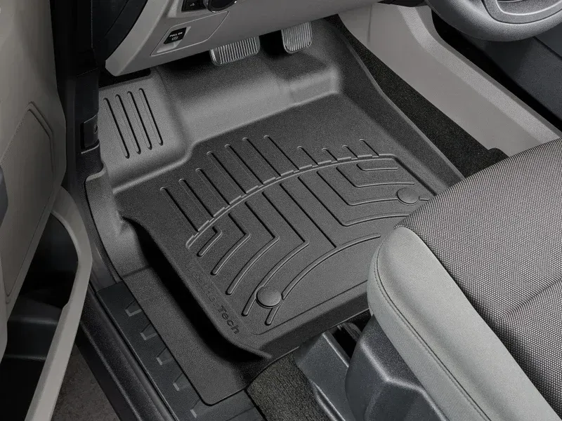 Car floor mat in a car’s front passenger footwell, black with raised ridges and gray trim.