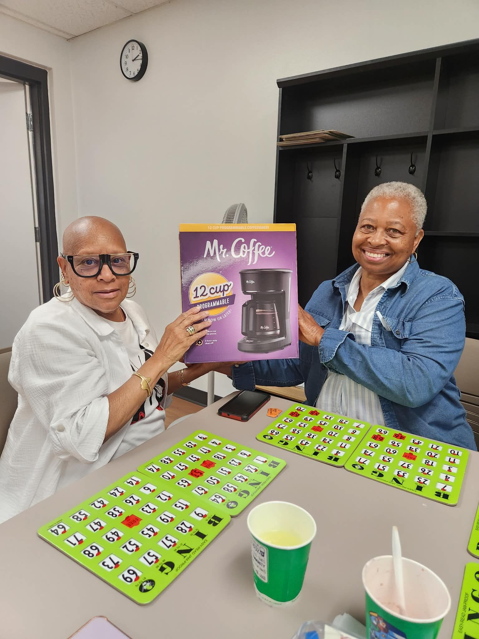 Two people smiling and holding a purple coffee maker box at a table with green game boards and cups.
