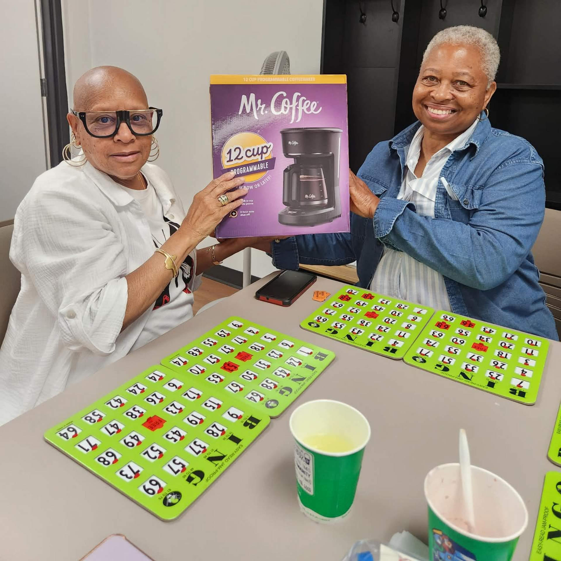Two people smiling and holding a purple coffee maker box at a table with green game boards and cups.