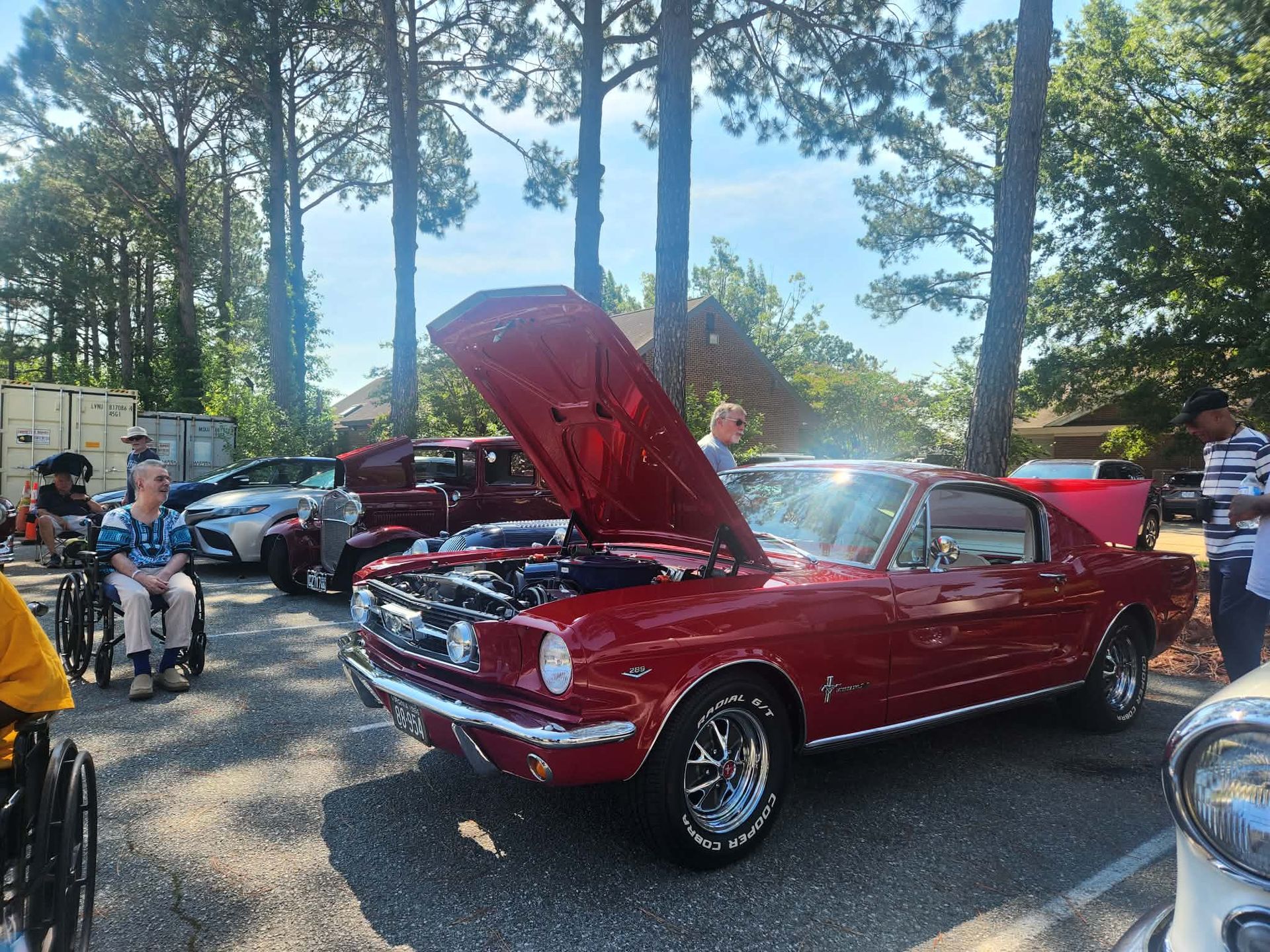 Red classic car with hood open at an outdoor car show among trees and other vehicles.