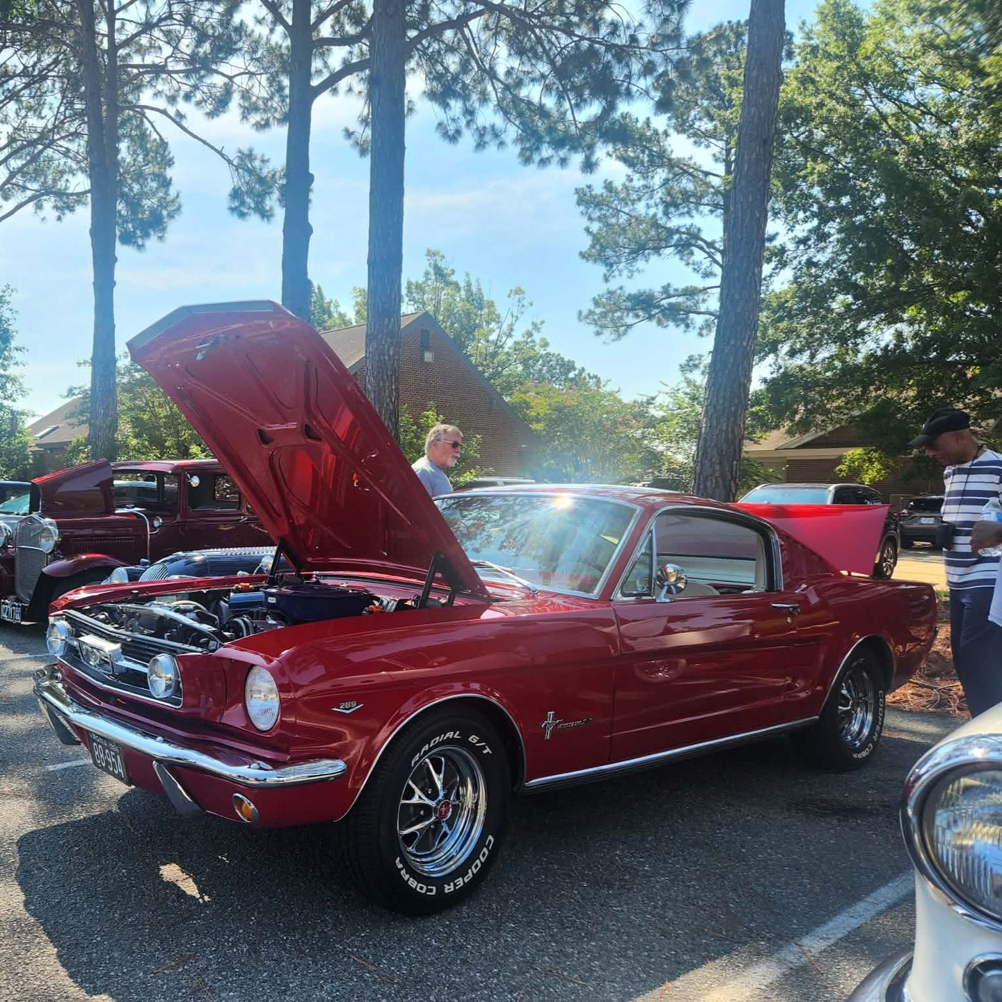 Red classic car with hood open at an outdoor car show among trees and other vehicles.