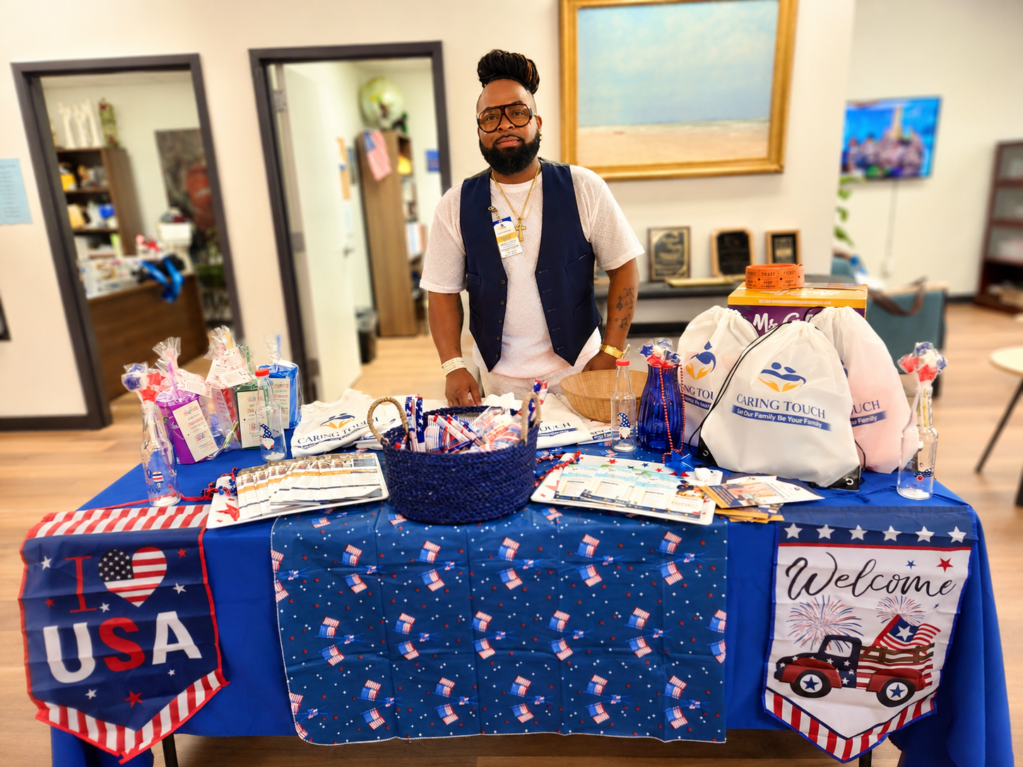 Man standing behind a blue patriotic-themed table with snacks and a Welcome sign, indoors.