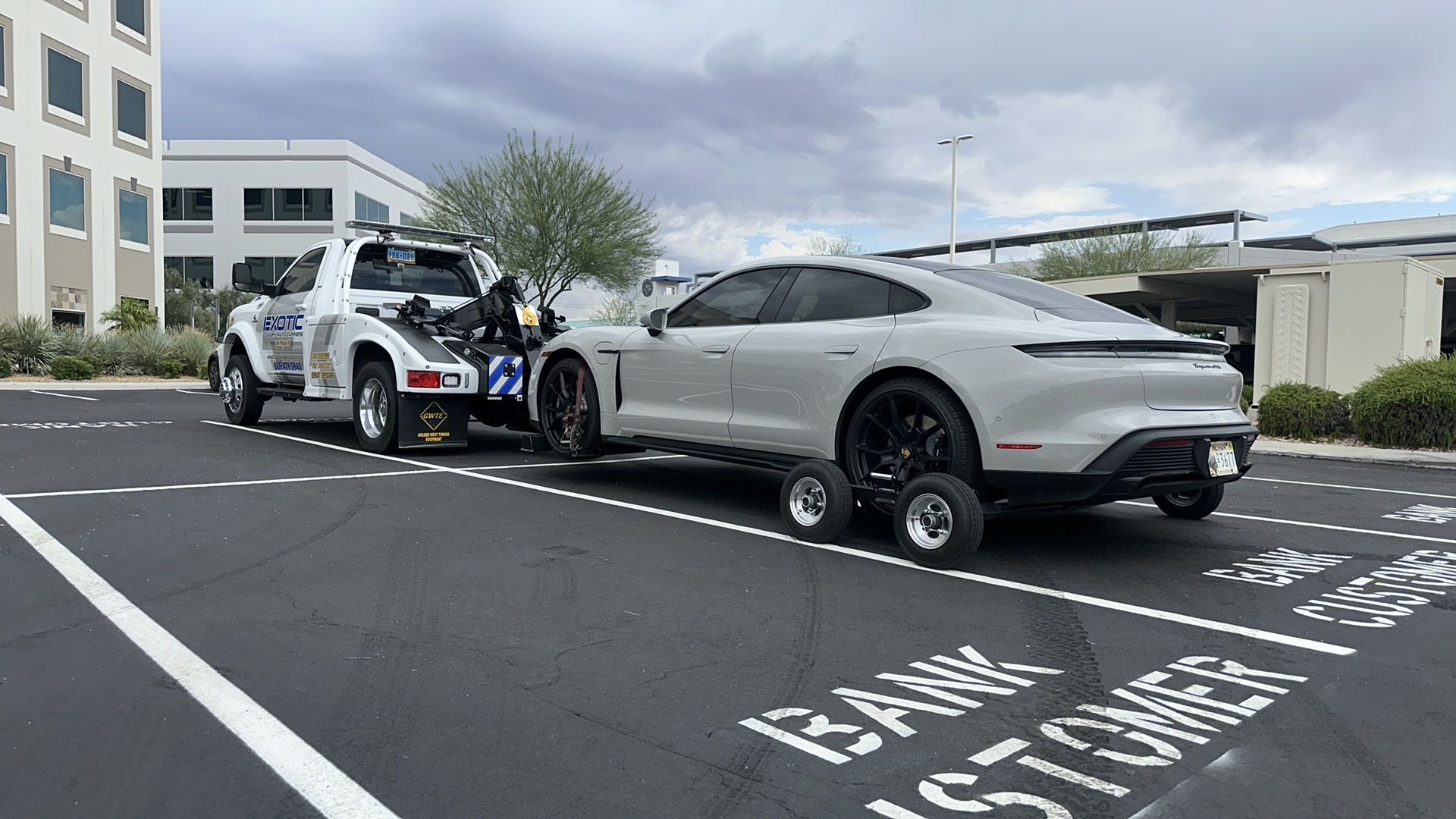 A light gray Porsche being towed by a white tow truck in a parking lot.