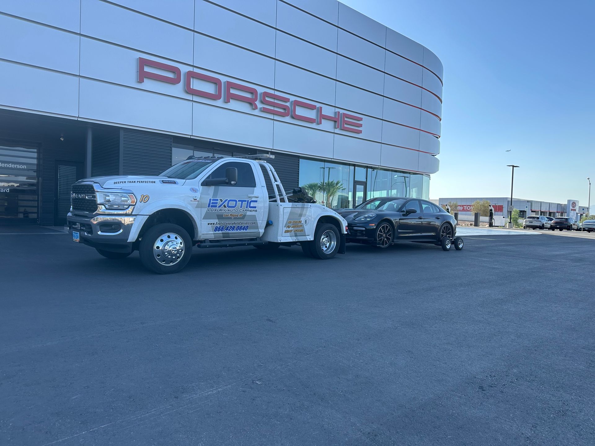 Dark gray car on trailer behind tow truck in front of Porsche building