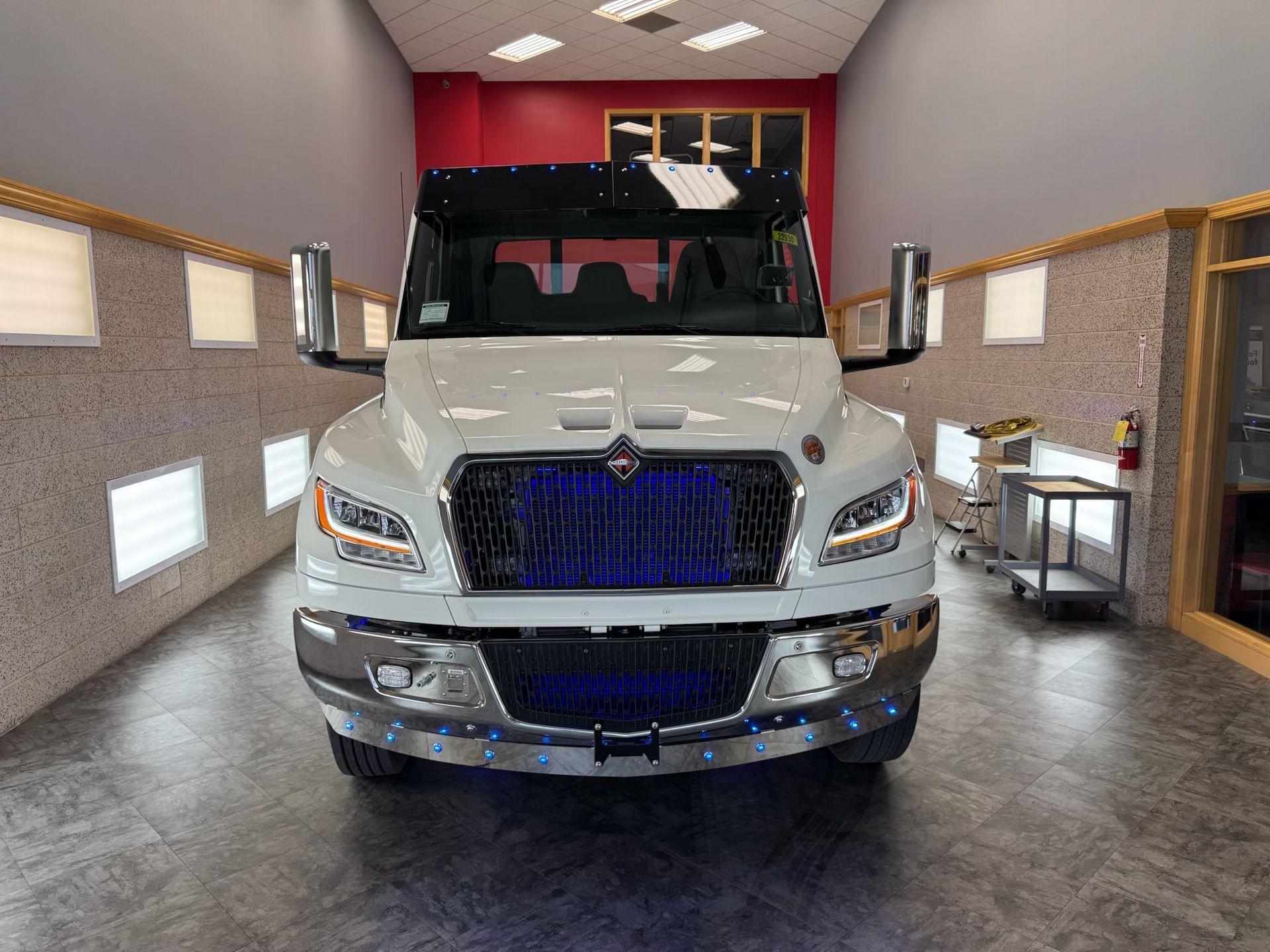 White semi-truck with blue grill lights, parked indoors, facing the camera. Chrome bumper, headlights on.
