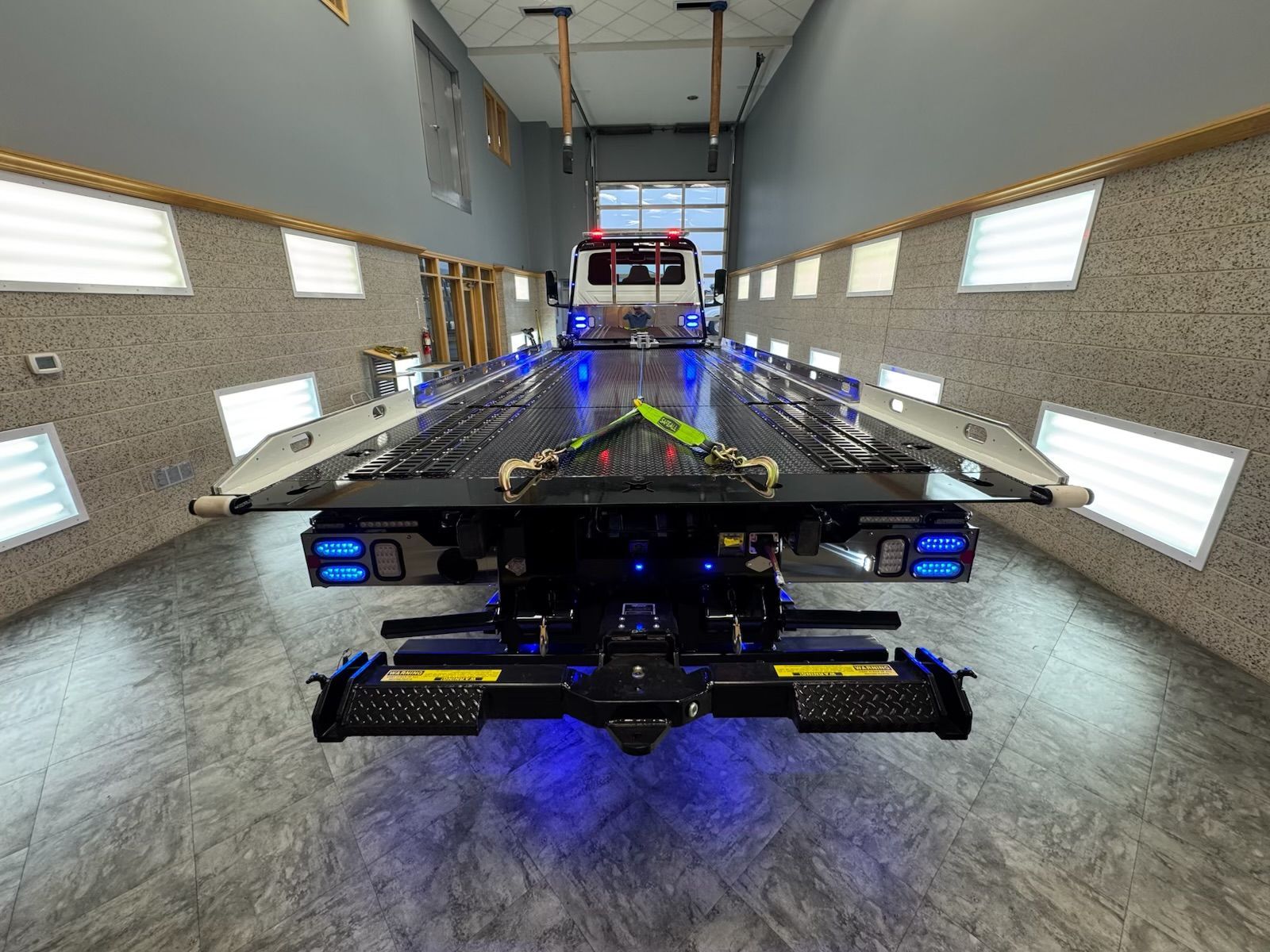 Tow truck bed with blue lights, parked in a gray garage with overhead lighting and a closed door in the distance.