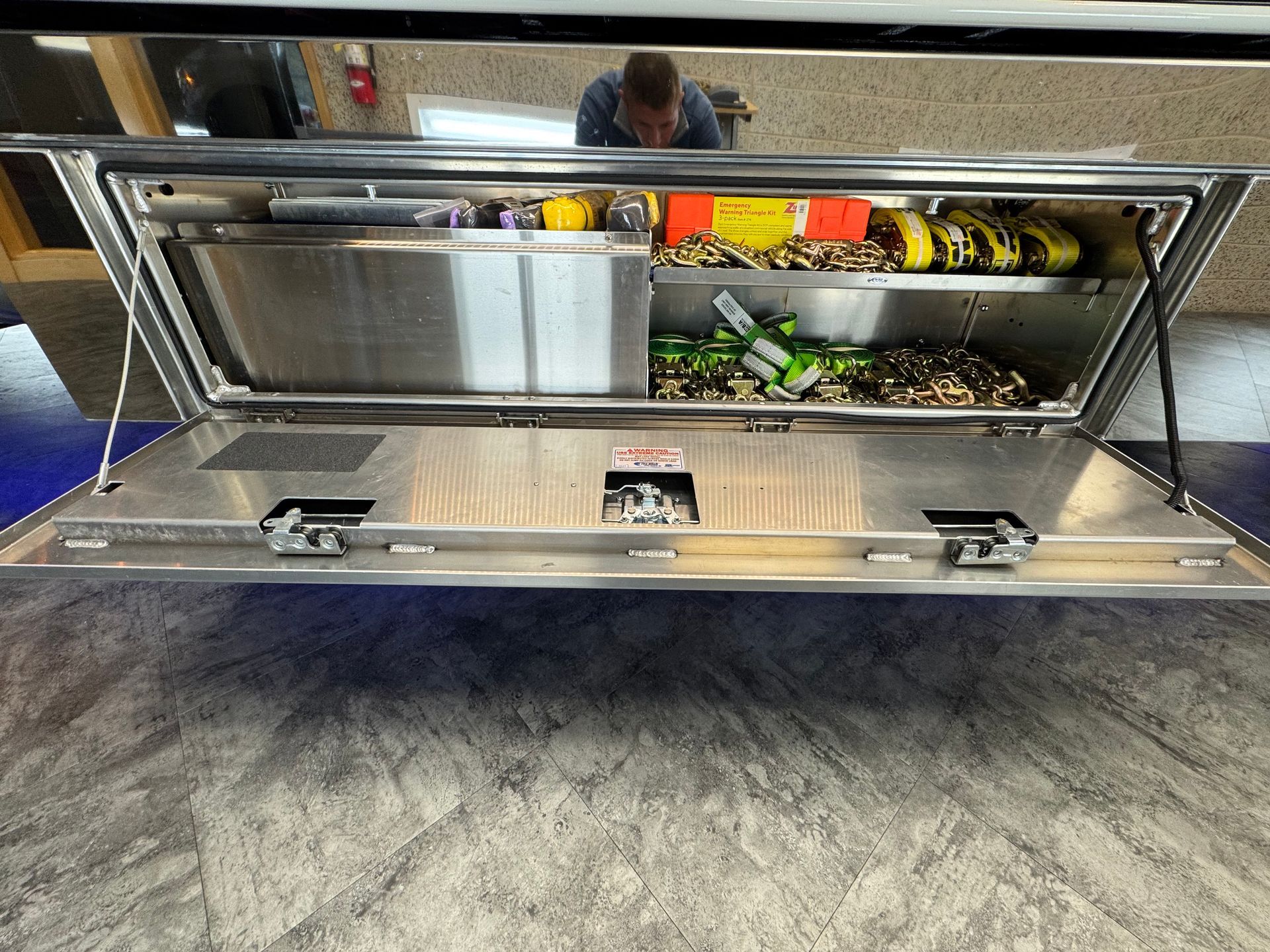 Open metal truck tool box with tools and person in the background.