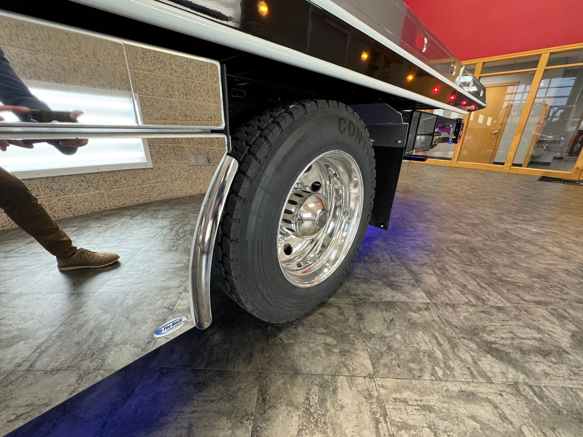 Shiny truck tire and fender; person's legs reflected in the chrome. Interior setting.