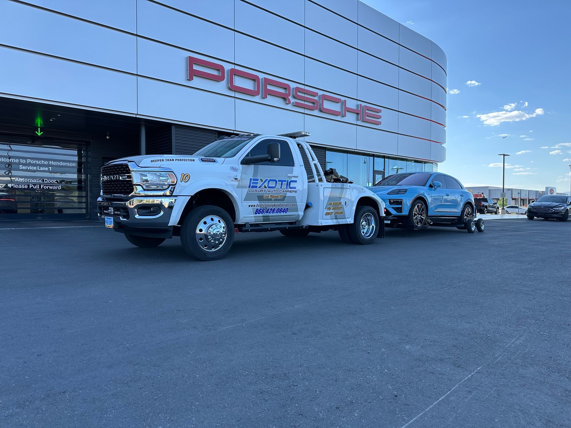 Tow truck towing a blue Porsche SUV in front of a Porsche dealership.