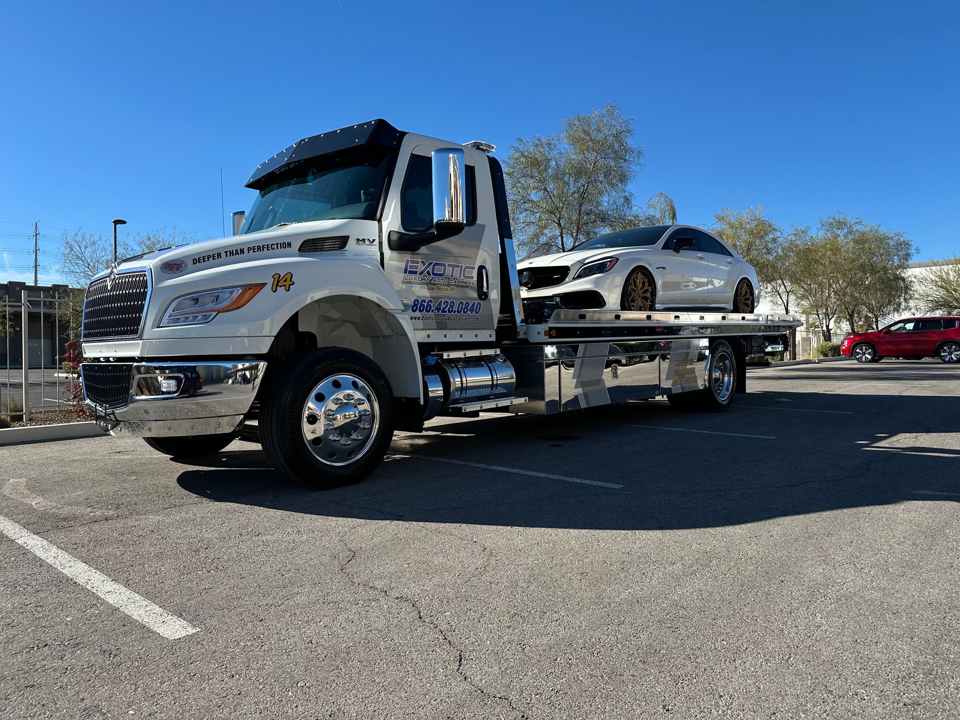 White tow truck with a white car on its flatbed, parked in a lot under a blue sky.