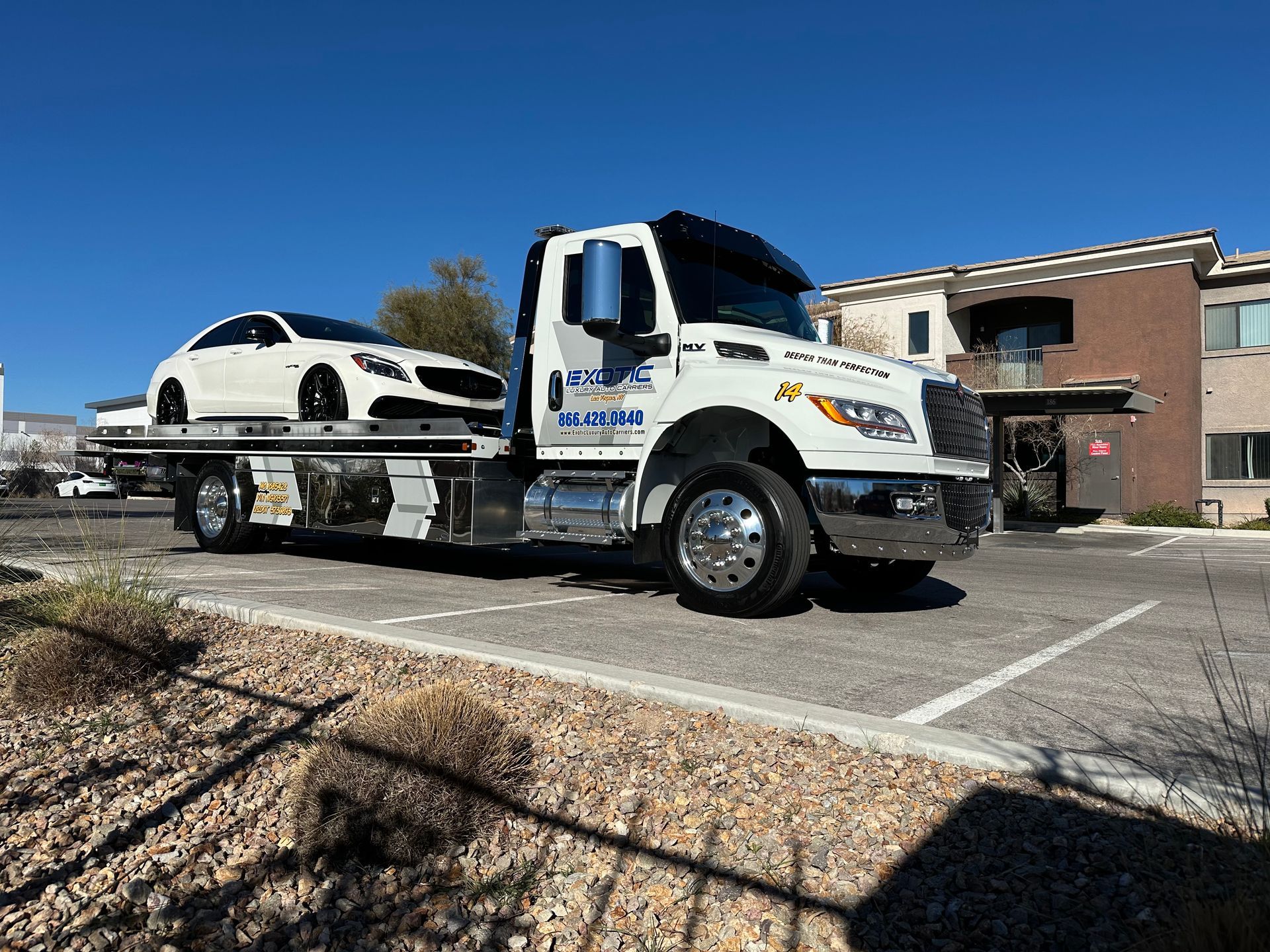 White tow truck with a white car on its flatbed in front of an apartment building on a sunny day.