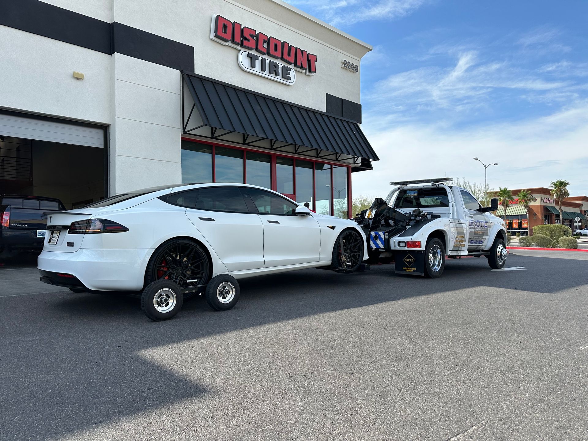 White Tesla being towed by a tow truck in front of a Discount Tire store on a sunny day.