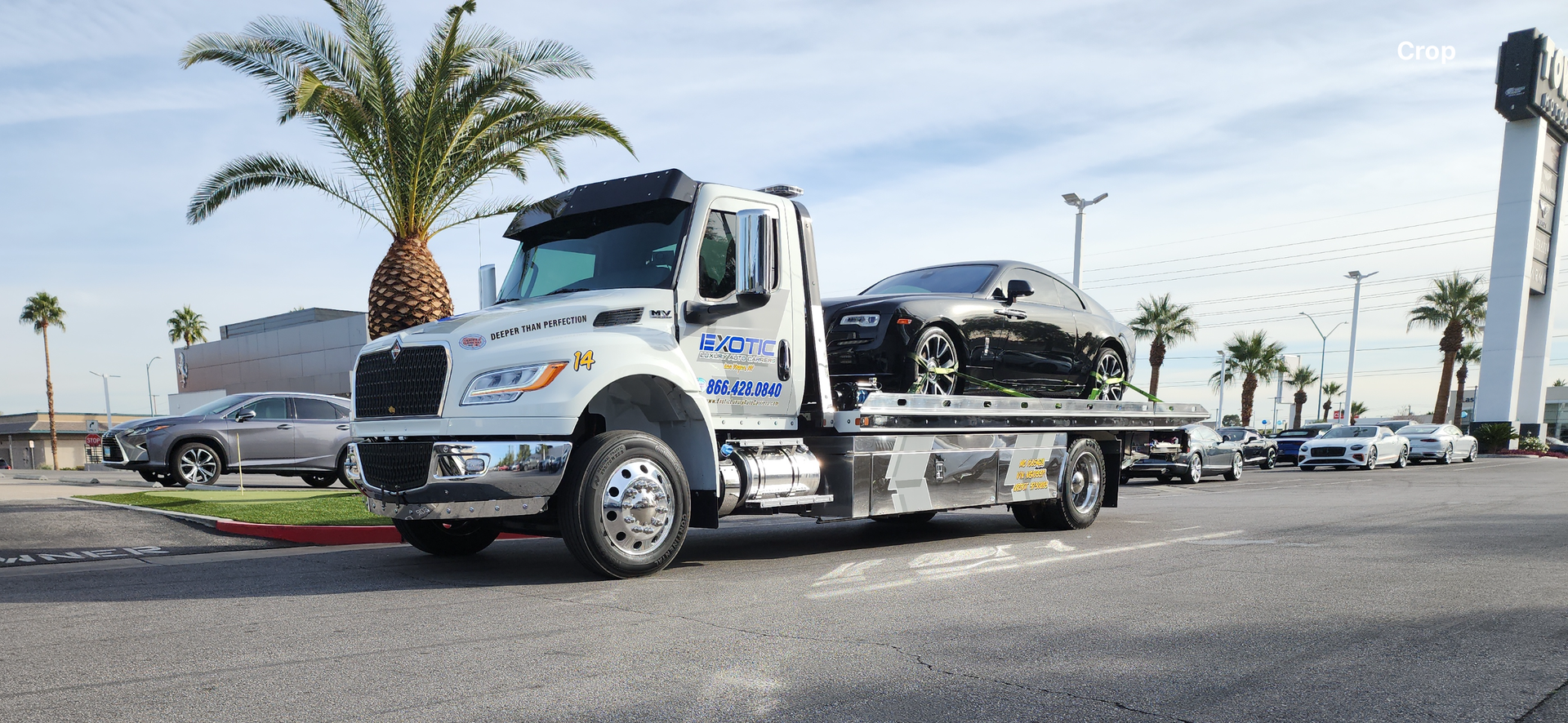 A white tow truck carrying a silver car at a dealership.