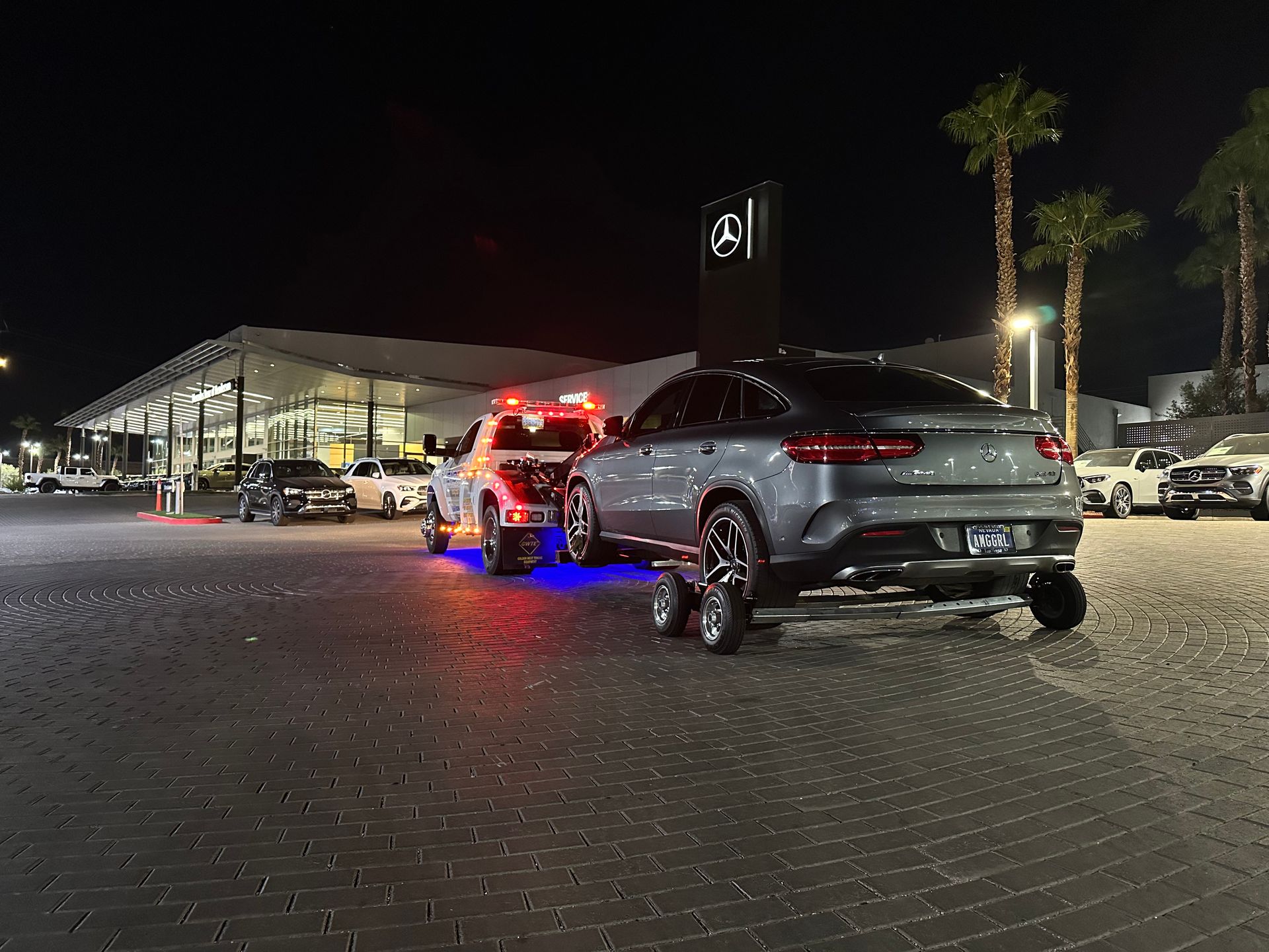 A gray Mercedes being towed at night in front of a Mercedes-Benz dealership.