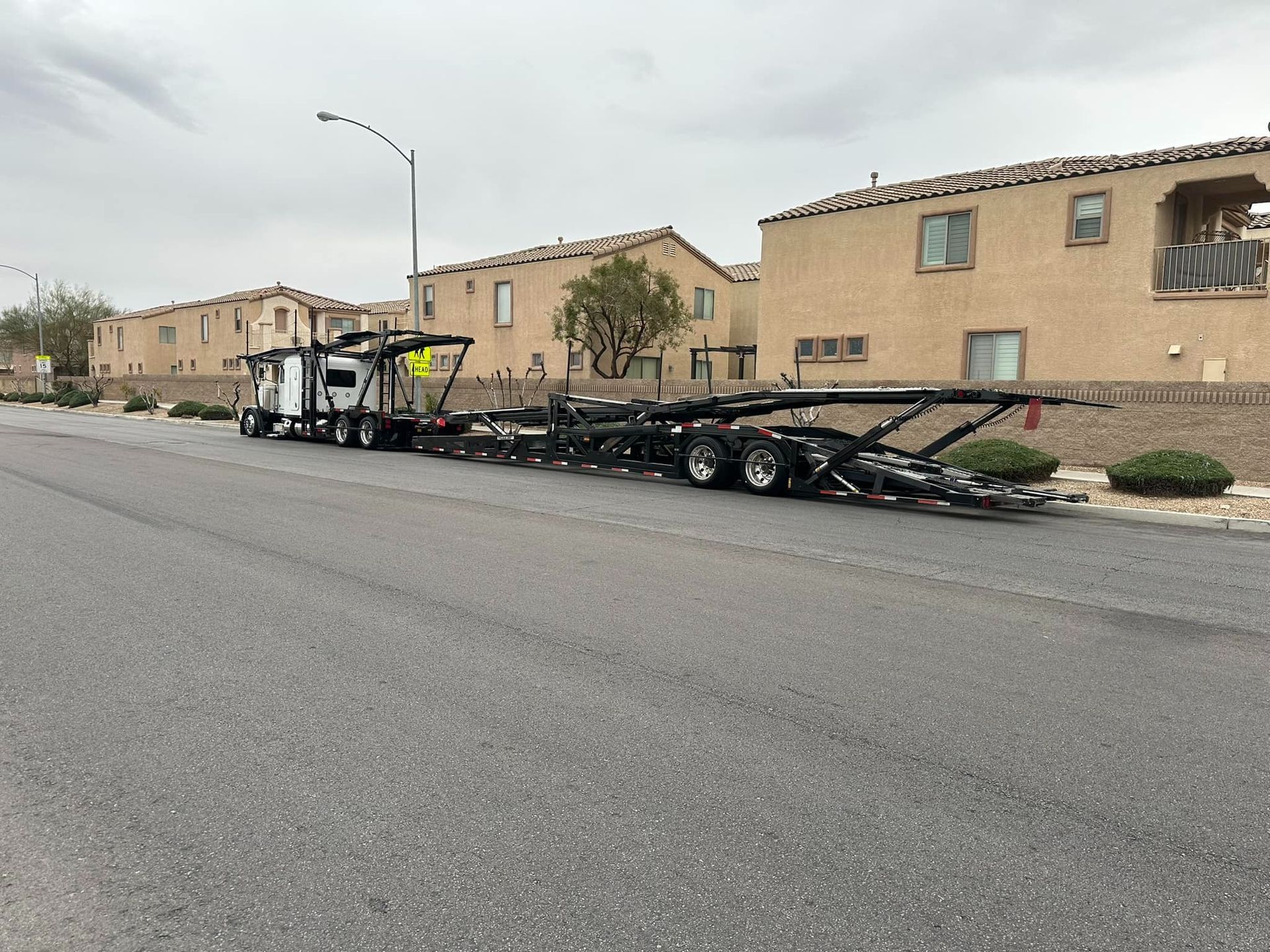 Car hauler truck parked on a residential street in front of two-story tan houses on a cloudy day.