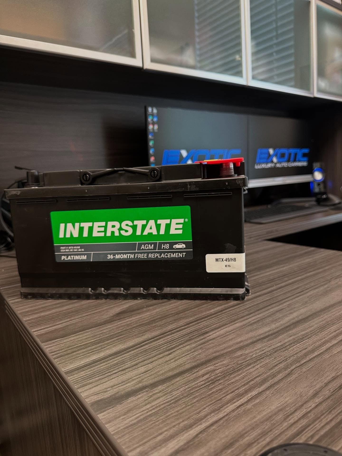 An Interstate car battery sits on a dark wood desk. The battery is black with a green label and red terminals.