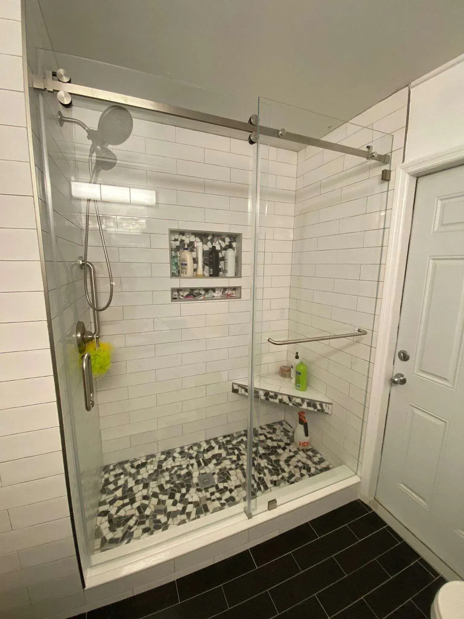 Glass shower with sliding door, white tile walls, black floor, and a shelf.
