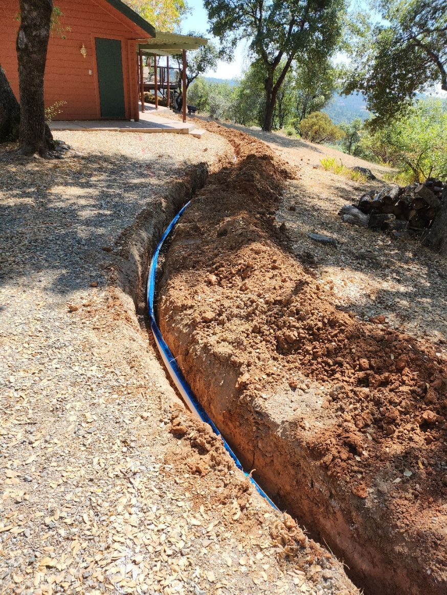 Trench dug in dirt with black pipe laid within. Small cabin and hillside in the background.