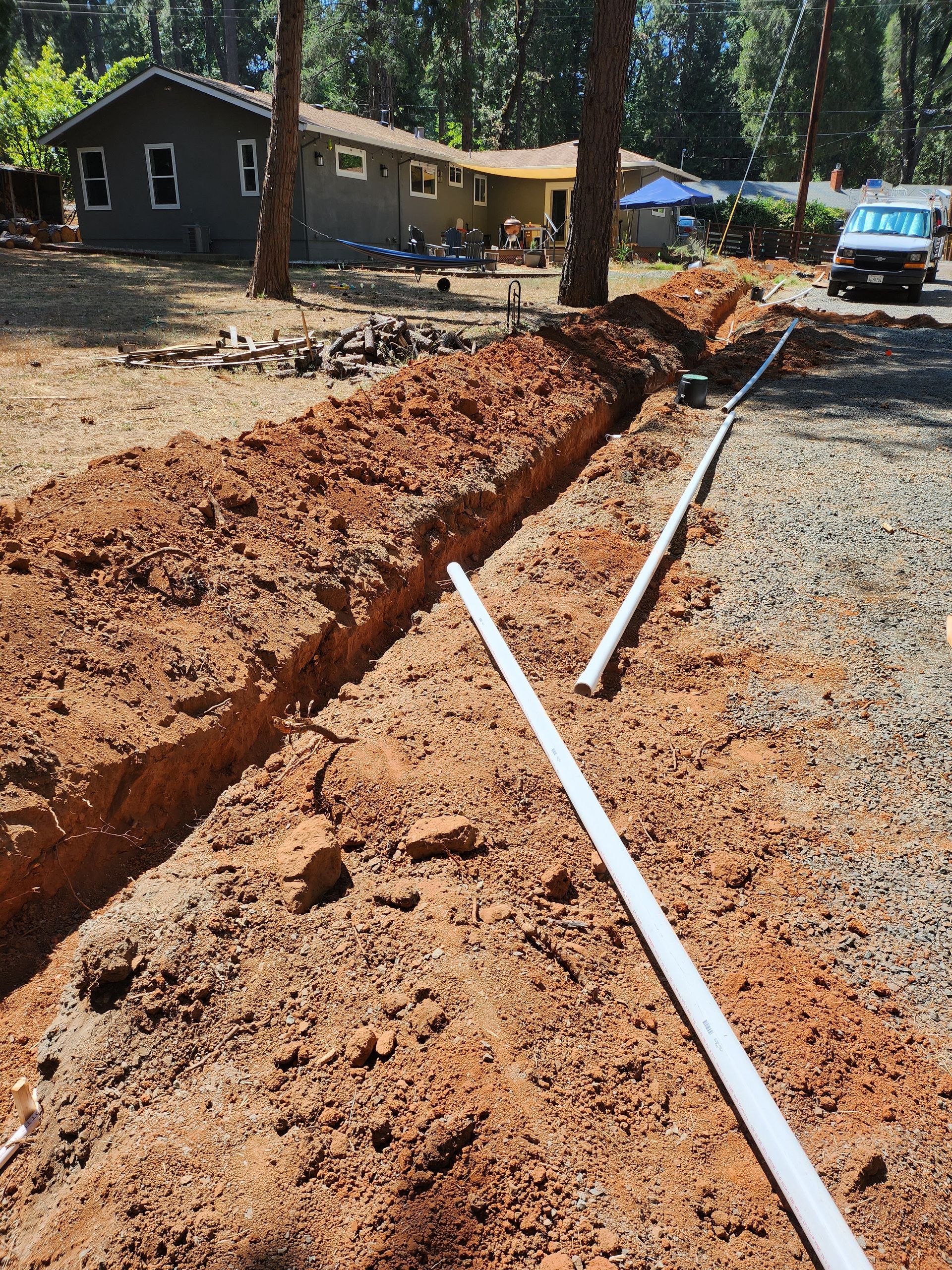 Trench dug in a yard with white pipes running through it, likely for utilities. House and vehicle visible in the background.