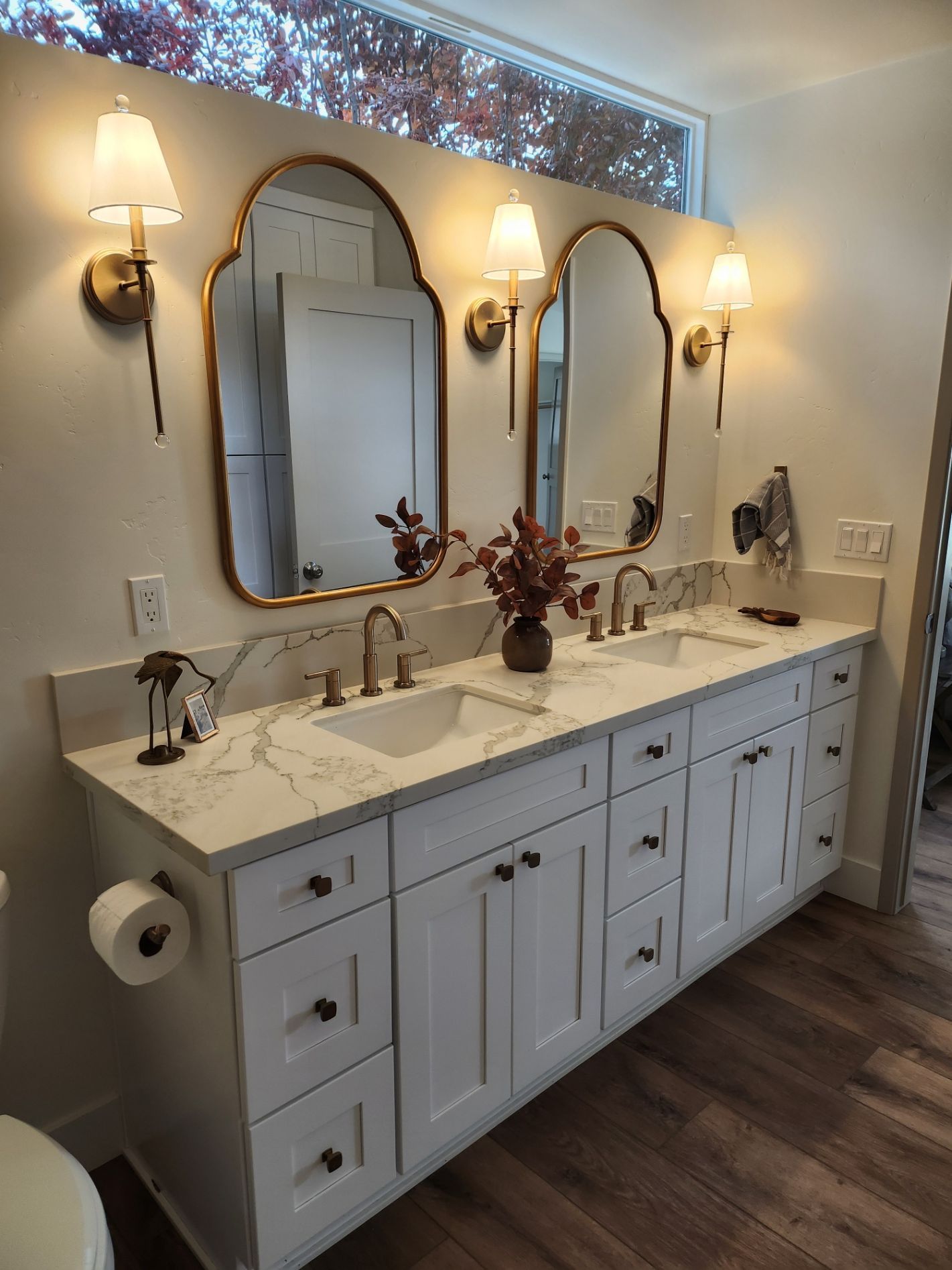 White bathroom vanity with two sinks, gold mirrors, sconces, and wooden floors.