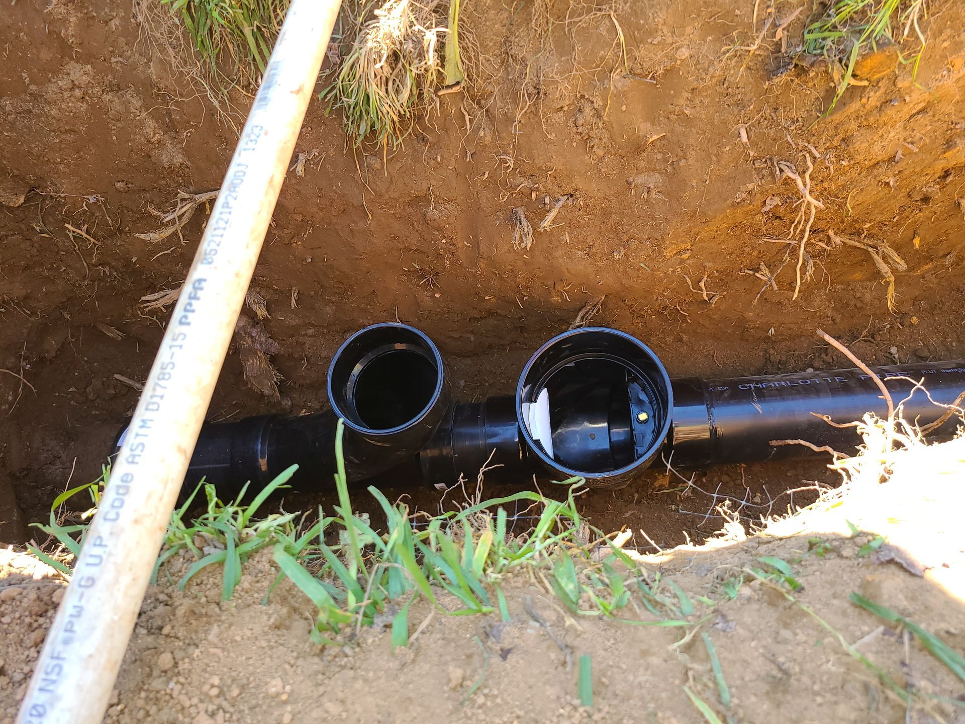 Black drainage pipes with two open ports in a dirt trench, near grass and a white PVC pipe.