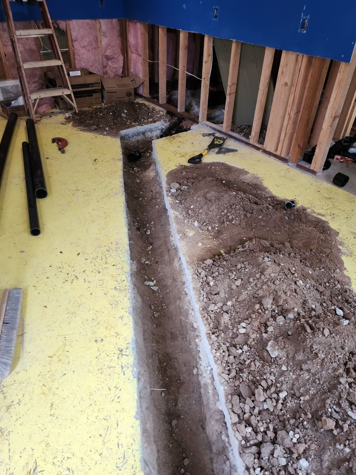 A trench dug in a room during construction. Dirt piles next to the trench. Yellow flooring, wooden studs.