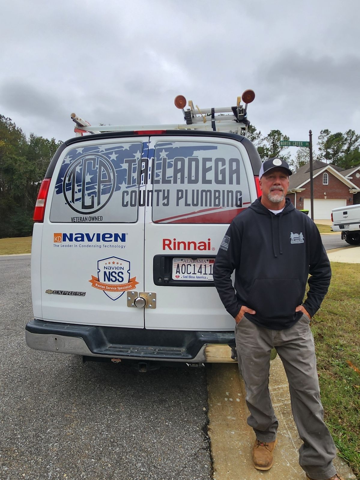 Man standing in front of a white plumbing van, with Talladega County Plumbing logo. Cloudy day, residential setting.